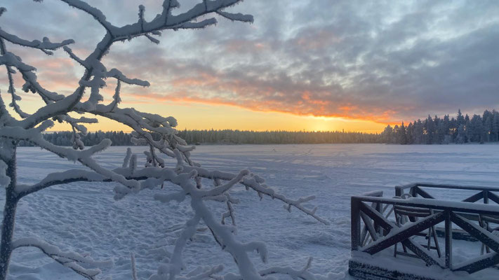 Sonnenuntergang über dem verschneiten See hinter Ästen und Holzgeländer